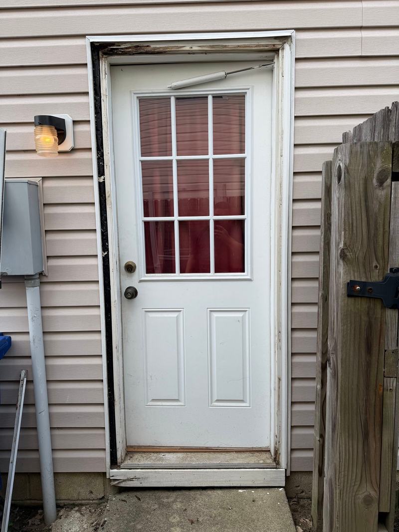 Older white exterior door with nine-lite window in tan vinyl siding beside a wooden gate