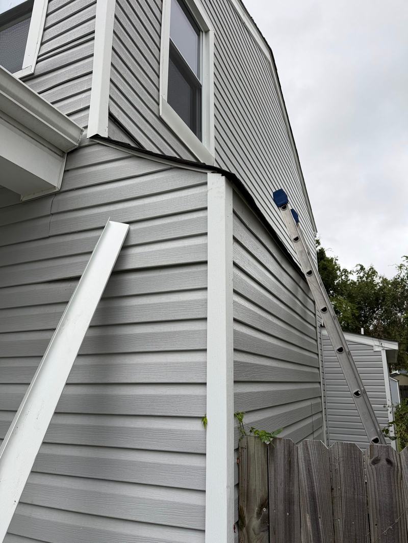 Gray vinyl siding corner with exposed overhang edge, loose white trim piece and a ladder