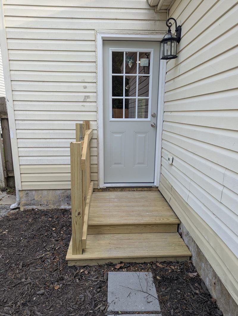 Wooden entry steps with handrail leading to a gray half-glass side door in yellow siding